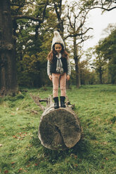 Little girl wearing cap standing on a tree trunk in autumnal park - CHAF000238