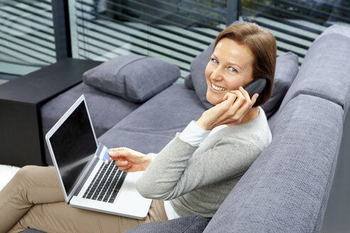 Porträt einer lächelnden Frau, die mit Laptop und Kreditkarte auf einer Couch sitzt und telefoniert - CHAF000270