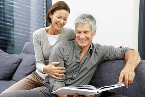 Porträt eines glücklichen Paares, das zusammen auf einer Couch sitzt und ein Buch betrachtet - CHAF000272