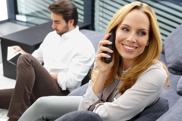 Portrait of smiling woman talking on telephone while her husband sitting in the background - CHAF000277