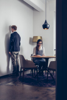 Junge Frau sitzt am Tisch mit einem Mann, der vor einer Wand steht - CHAF000305