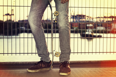 Germany, Hamburg, legs of a teenage boy with skateboard leaning against a fence - MEMF000808