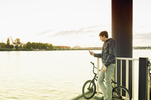 Deutschland, Hamburg, Jugendlicher mit BMX auf einem Steg stehend und mit Smartphone - MEMF000813