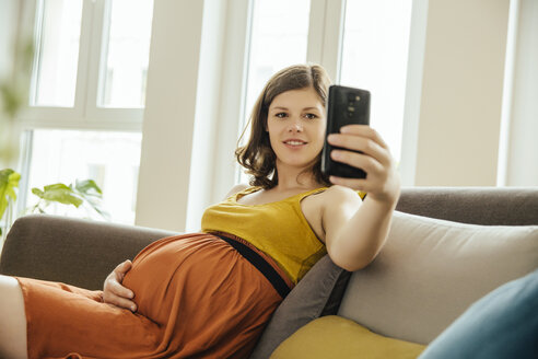 Schwangere Frau macht ein Selfie mit Smartphone auf ihrer Couch zu Hause - MFF001786