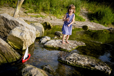 Kleines Mädchen spielt mit Holzspielzeugboot am Flussufer, lizenzfreies Stockfoto