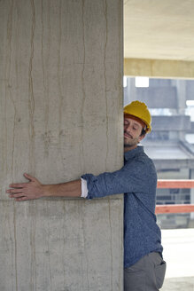 Man with hard hat embracing concrete pillar on construction site - FMKF001583