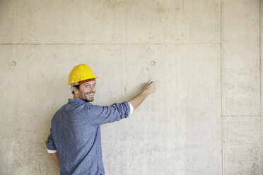 Smiling man with hard hat on construction site drawing on concrete wall - FMKF001629