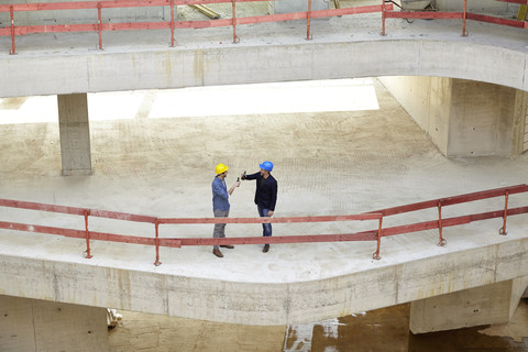 Zwei Männer mit Schutzhelmen stoßen auf einer Baustelle mit Bierflaschen an, lizenzfreies Stockfoto
