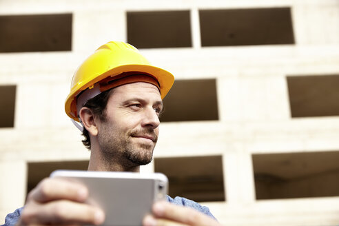 Mann mit Schutzhelm auf einer Baustelle, der ein Mobiltelefon hält - FMKF001634