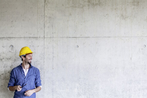 Lächelnder Mann mit Schutzhelm auf einer Baustelle an einer Betonwand - FMKF001730