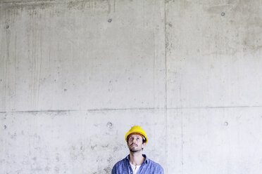 Mann mit Schutzhelm auf einer Baustelle an einer Betonwand mit Blick nach oben - FMKF001660