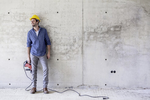 Lächelnder Mann mit Schutzhelm auf einer Baustelle mit Bohrmaschine - FMKF001664