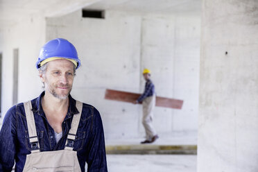 Lächelnder Arbeiter auf einer Baustelle mit einem Kollegen im Hintergrund - FMKF001677