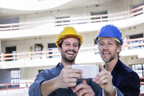 Zwei glückliche Männer auf der Baustelle schauen auf ihr Handy - FMKF001699