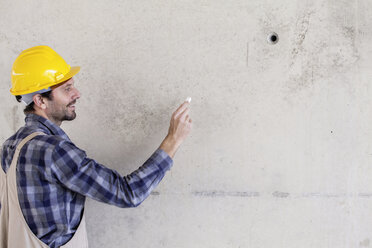 Mann mit Schutzhelm auf einer Baustelle, der auf eine Betonwand zeichnet - FMKF001710