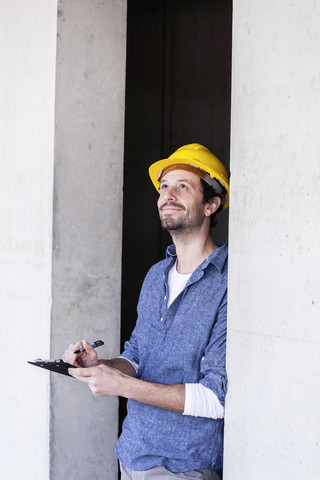 Mann mit Schutzhelm auf einer Baustelle, der nach oben schaut und auf ein Klemmbrett schreibt, lizenzfreies Stockfoto