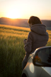 Germany, woman leaning on car in front of a field at sunrise - MIDF000493