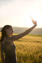 Germany, woman standing in front of a field at sunrise taking a selfie with her smartphone - MIDF000500