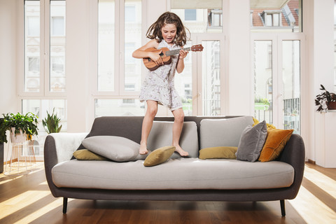 Glückliches Mädchen spielt Mini-Gitarre beim Springen auf einer Couch im Wohnzimmer zu Hause, lizenzfreies Stockfoto