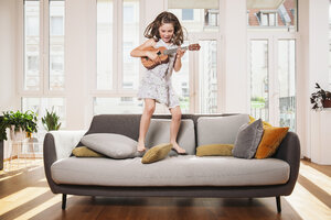 Glückliches Mädchen spielt Mini-Gitarre beim Springen auf einer Couch im Wohnzimmer zu Hause - MFF001692