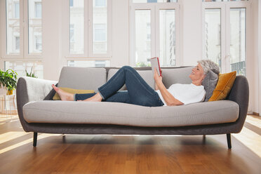 Ältere Frau entspannt sich mit digitalem Tablet auf einer Couch im Wohnzimmer - MFF001746