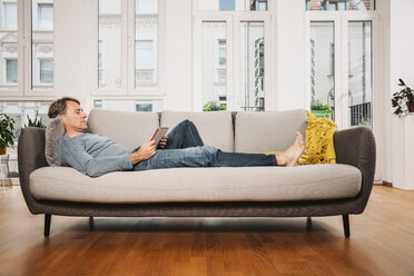 Man relaxing with digital tablet on a couch at living room - MFF001748