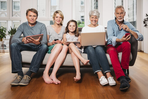 Group picture of three generations family with different digital devices sitting on one couch - MFF001695
