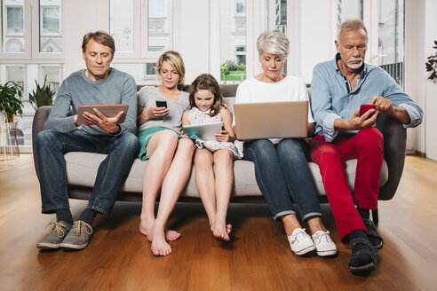 Gruppenbild einer Familie mit drei Generationen, die auf einer Couch sitzt und verschiedene digitale Geräte benutzt - MFF001696