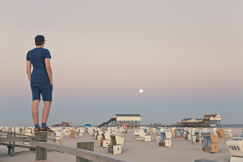 Germany, St Peter-Ording, young man standing on wooden fence looking at the beach stock photo