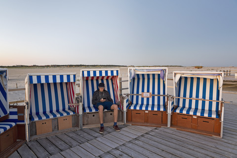 Germany, St Peter-Ording, teenage boy with smartphone sitting in hooded beach chair stock photo