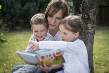 Mutter und ihre beiden Kinder sitzen auf einer Wiese und lesen ein Bilderbuch - PAF001464