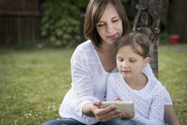 Mutter und ihre kleine Tochter sitzen auf einer Wiese und schauen auf ein Smartphone - PAF001460
