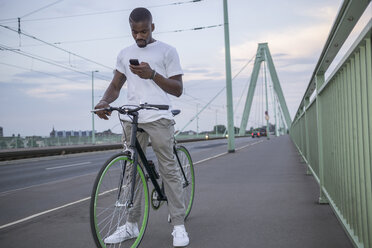 Deutschland, Köln, junger Mann steht mit Fahrrad auf der Rheinbrücke und schaut auf sein Smartphone - RIBF000152