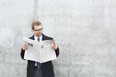 Businessman reading newspaper at concrete wall - WESTF021321