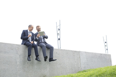 Zwei Geschäftsleute mit digitalem Tablet sitzen an einer Betonwand, lizenzfreies Stockfoto