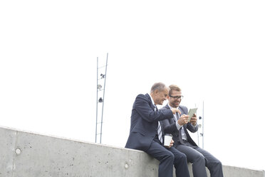 Two businessmen with digital tablet sitting on concrete wall - WESTF021309