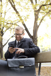 Smiling businessman on park bench looking at cell phone - WESTF021361