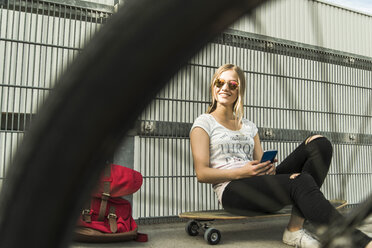 Teenager-Mädchen sitzt auf einem Skateboard und hält ein Mobiltelefon in der Hand - UUF004885