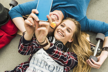 Happy young couple lying on ground with skateboard looking at cell phone - UUF004899