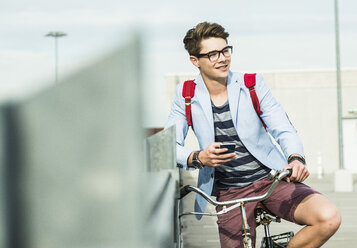 Lächelnder junger Mann mit Fahrrad und Mobiltelefon auf dem Parkdeck - UUF004905