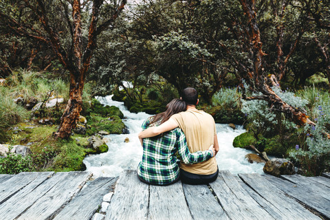 Peru, Paar sitzt Arm in Arm auf einer Holzbrücke und genießt die Natur, lizenzfreies Stockfoto