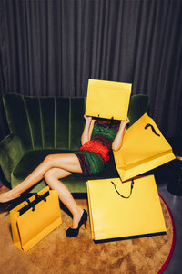 Junge Frau sitzt auf einer Couch und versteckt ihr Gesicht hinter einer Einkaufstasche - CHAF000535