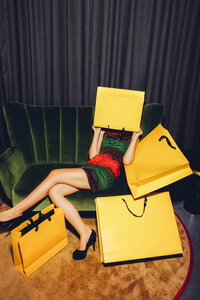 Young woman sitting on couch hiding her face behind a shopping bag - CHAF000535