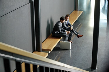 Zwei junge Geschäftsleute sitzen auf einer Bank und benutzen ein Mobiltelefon in einer Büro-Lobby - CHAF000395