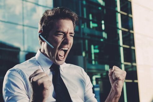 Excited businessman with clenched fists screaming outside office building - CHAF000413