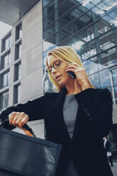 Geschäftsfrau, die die Zeit prüft, während sie vor einem Bürogebäude mit einem Handy telefoniert - CHAF000547