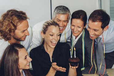 Excited young businesswoman holding a small muffin with burning candles celebrating her birthday in office with colleagues - CHAF000485