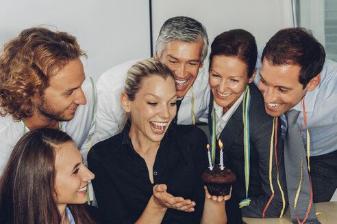 Aufgeregte junge Geschäftsfrau, die einen kleinen Muffin mit brennenden Kerzen hält und ihren Geburtstag im Büro mit Kollegen feiert - CHAF000485