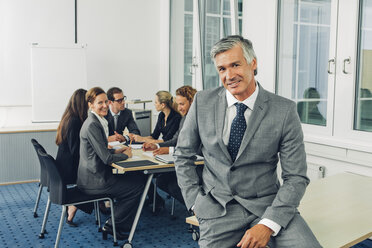 Mature businessman sitting on desk, team working in background - CHAF000508