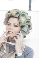 Portrait of woman with hair curlers smoking a cigarette while talking on phone - ZEF006296